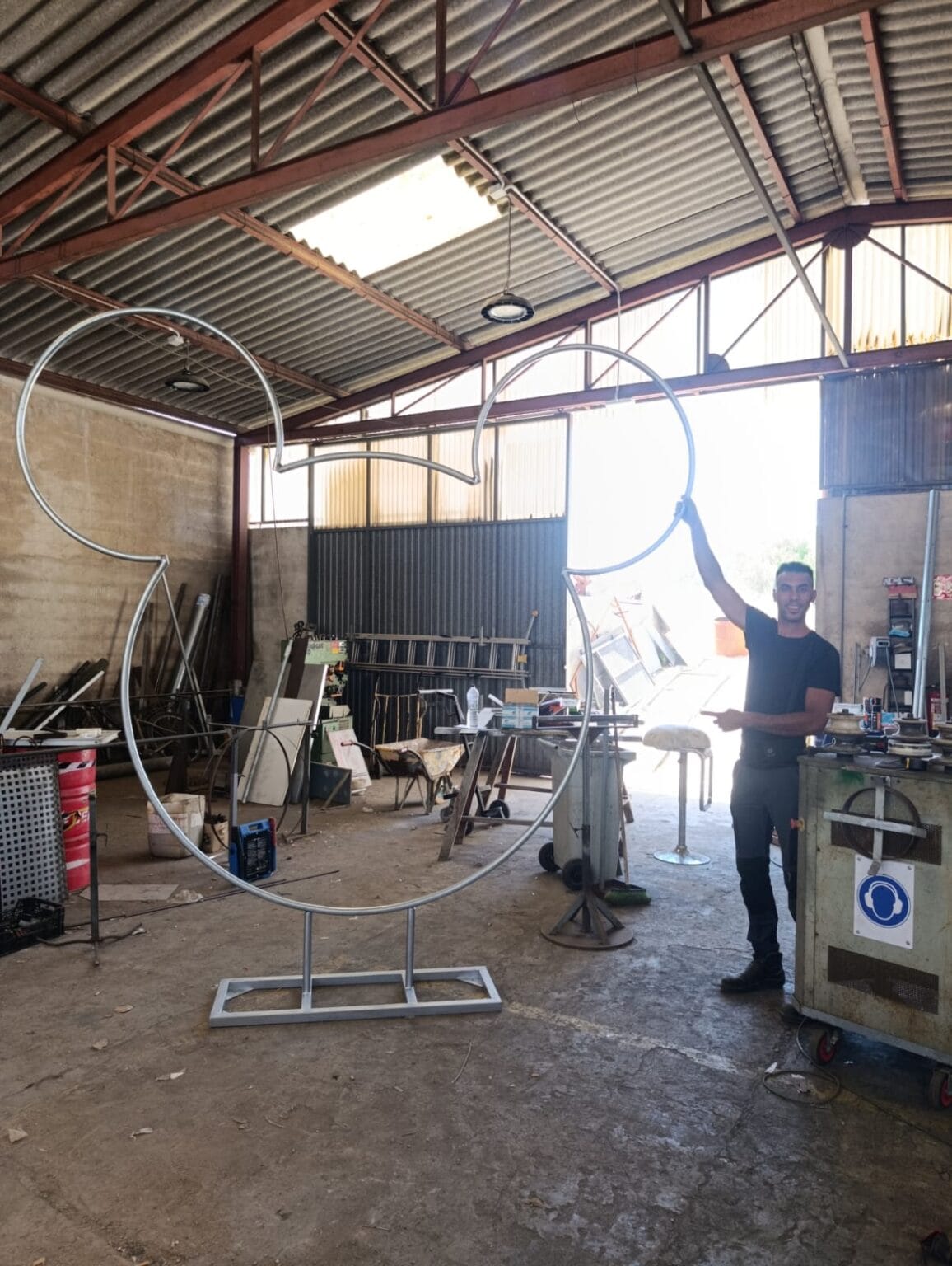 Una persona se encuentra en un taller junto a una gran escultura metálica abstracta con forma de curva fluida. El taller está lleno de herramientas, equipos y materiales metálicos, y la luz del sol entra a raudales por la puerta abierta.